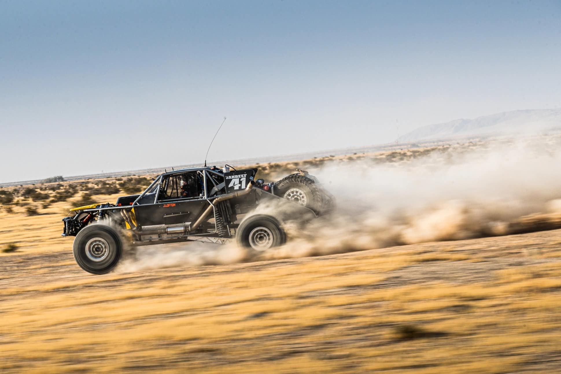 A racing truck driving across dirt terrain throwing dust behind it.